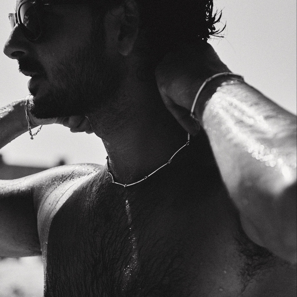 Black and white photo of a person with sunglasses and a necklace, possibly on a beach.