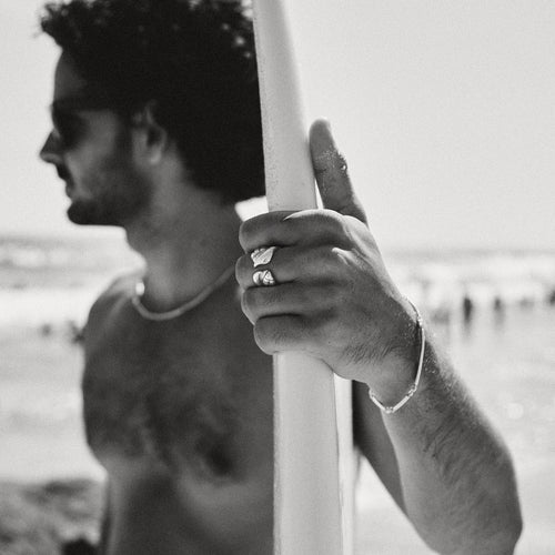 A man holding a surfboard on a beach with a blurred background, wearing a silver jewelry ring and bracelet.