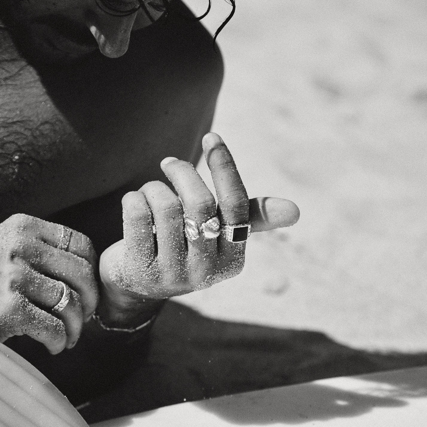 Person sitting on a beach with a surfboard, wearing a ring.