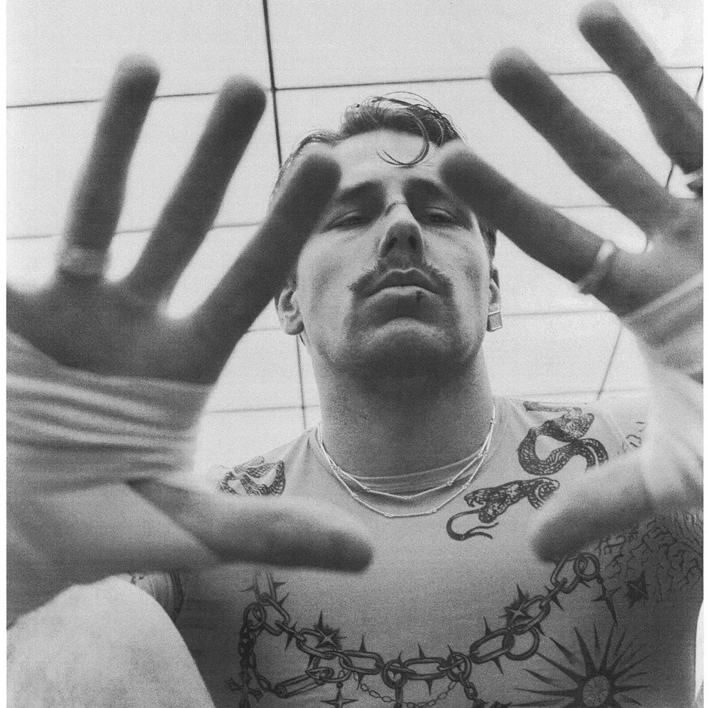 Man with tattoos on his arms and chest, wearing a white tank top, with a tiled ceiling in the background.
