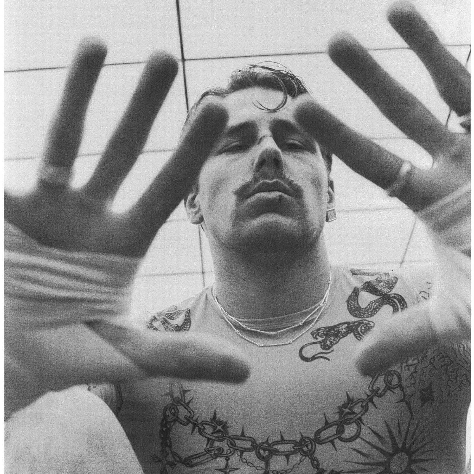 Man with tattoos on his arms and chest, wearing a white tank top, with a tiled ceiling in the background.