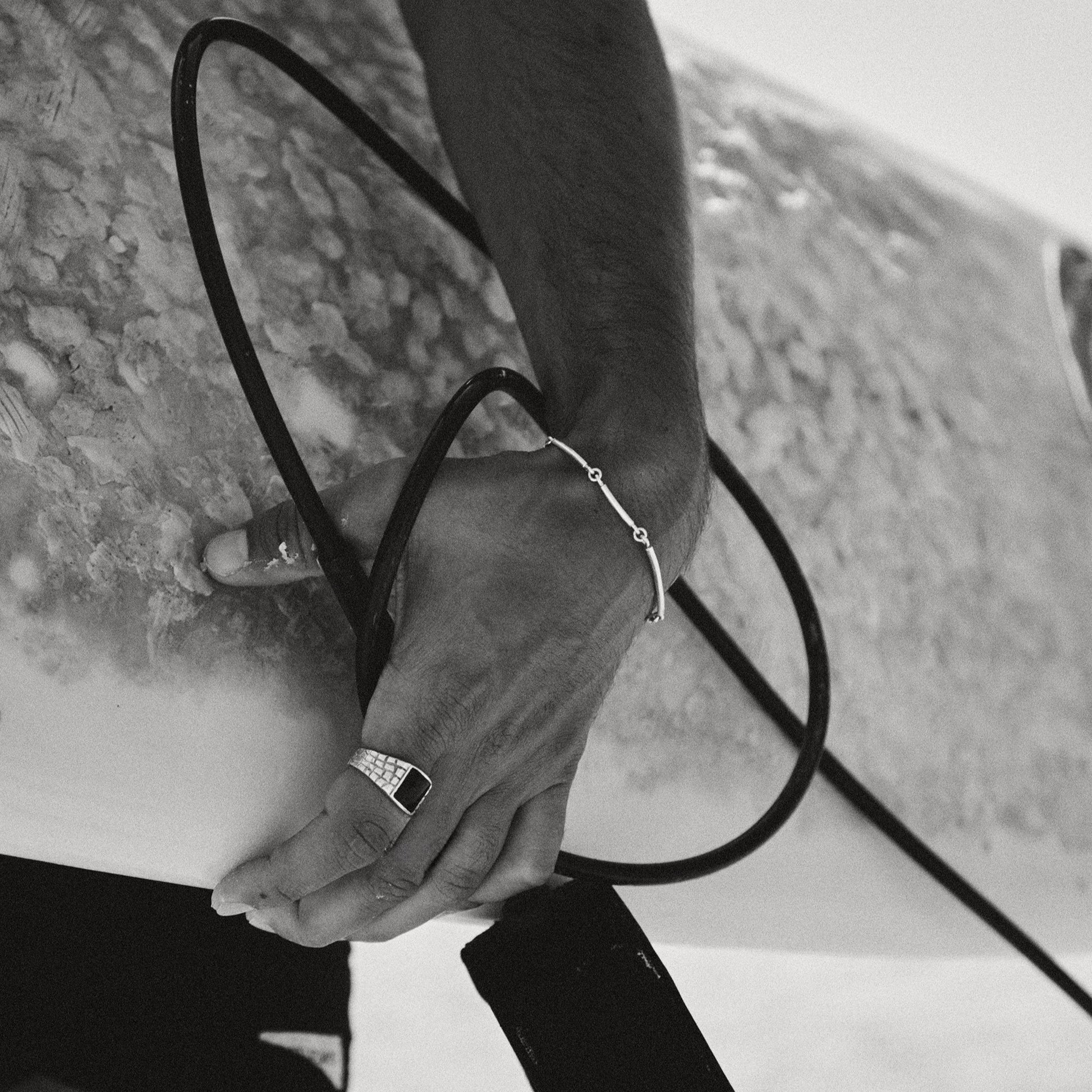 Person holding a surfboard with a close-up of their hand wearing a bracelet and ring.