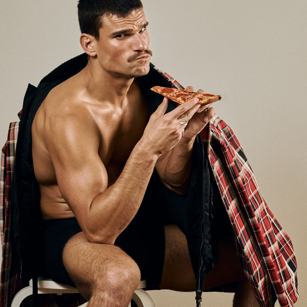 Man sitting on a chair eating pizza with a shirt draped over his shoulders.