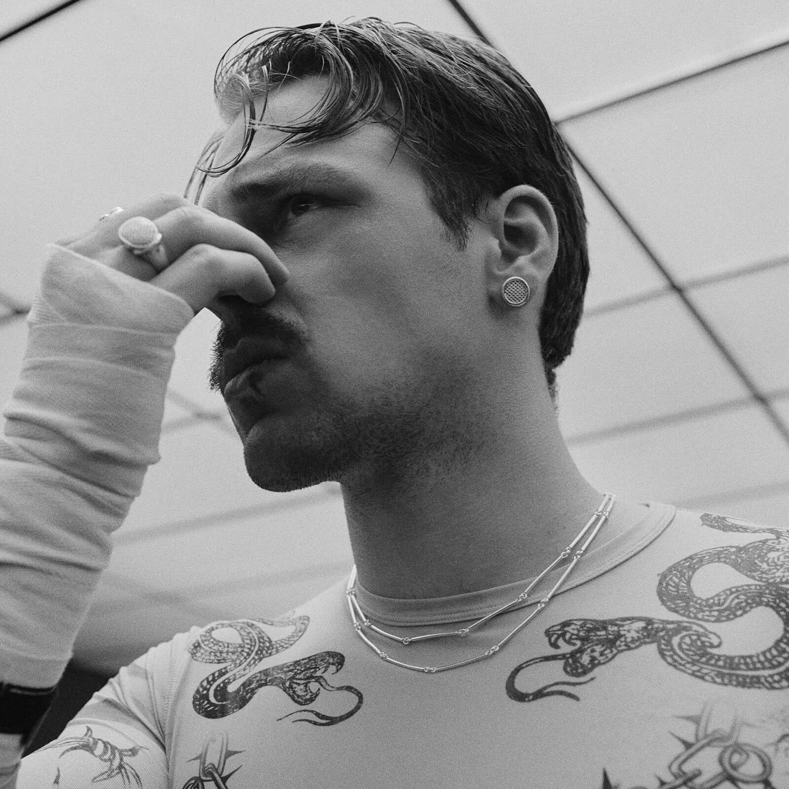 Man with tattoos on his arms wearing a white shirt against a geometric-patterned wall.